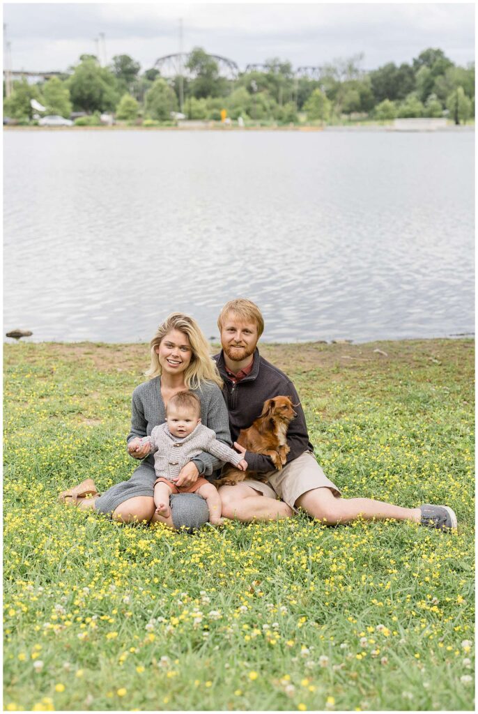 New parents of 6 month old son and their Dachsund dog sit together in front of a lake at Shelby Bottom Nature Preserve during their Nashville family session with Wisp + Willow Photography Co..
