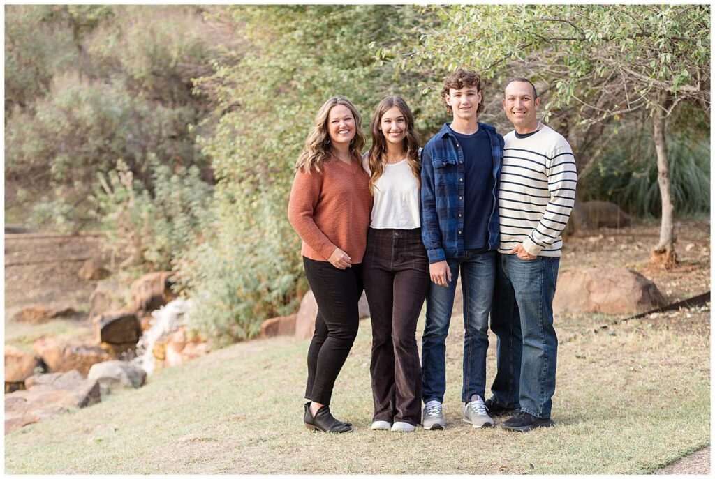 A family of 4 with a teenage son and daughter, stand together and smile at the camera of Wisp + Willow Photography Co. during their Frisco mini session.  They all wear pants and orange, white, and blue coordinating colors.