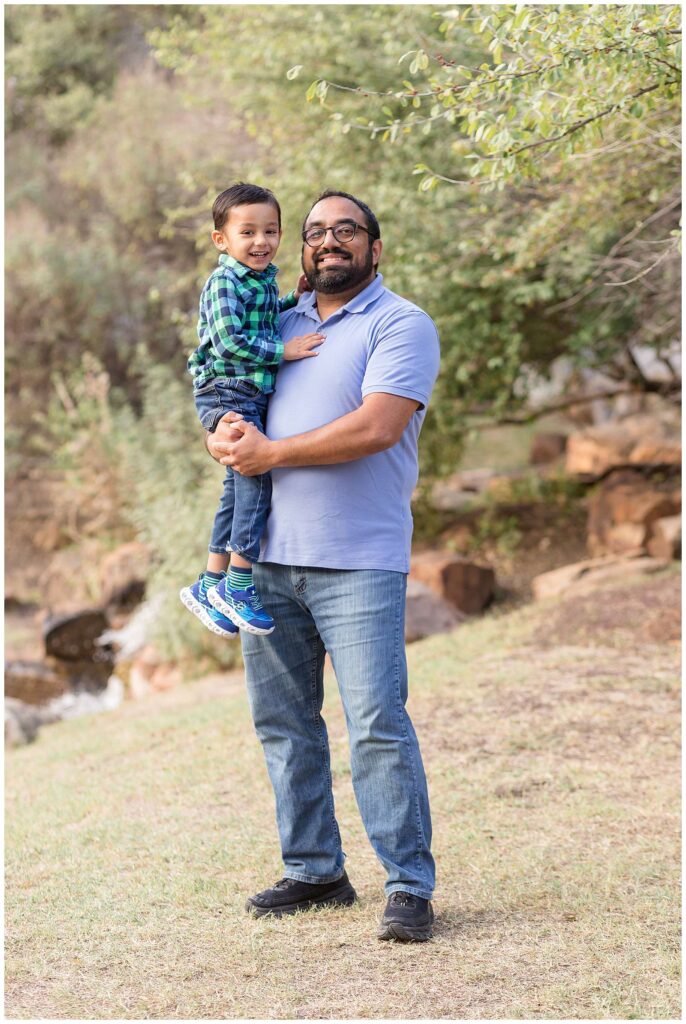 Preschool aged son and Dad smile at the camera during their fall mini sessions.  Dad holds son and they coordinate by Dad wearing a blue shirt and jeans and his son wears a green and blue plaid shirt and jeans.