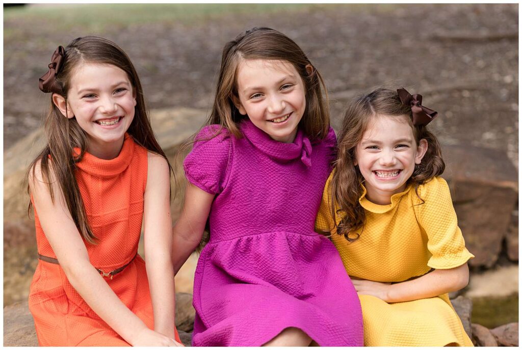 3 sisters smile big as they sit on some rocks at Frisco Central Park with one wearing an orange sweater, a pink/purple dress, and the last wearing a yellow dress.