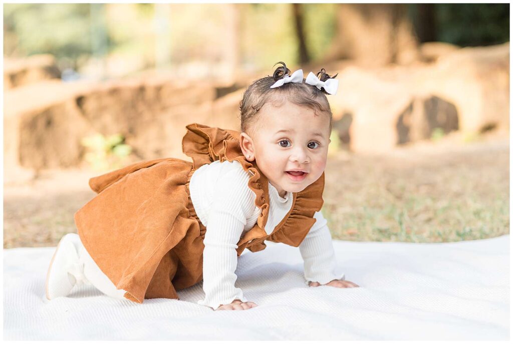 A sweet baby is up on all fours on a white blanket and wears white tights, a long sleeve white shirt and shoes, and a tan/brown suede colored dress during Frisco mini sessions.