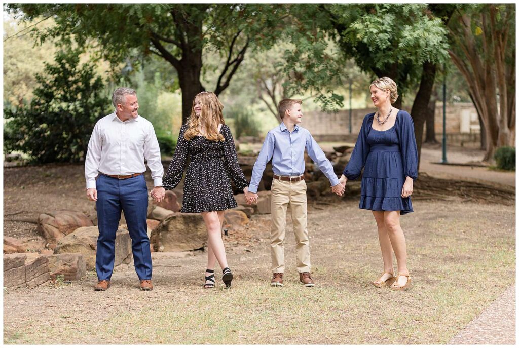 Family of 4, with a teenage son and daughter, all hold hands and look and smile at each other during their Frisco  mini session.