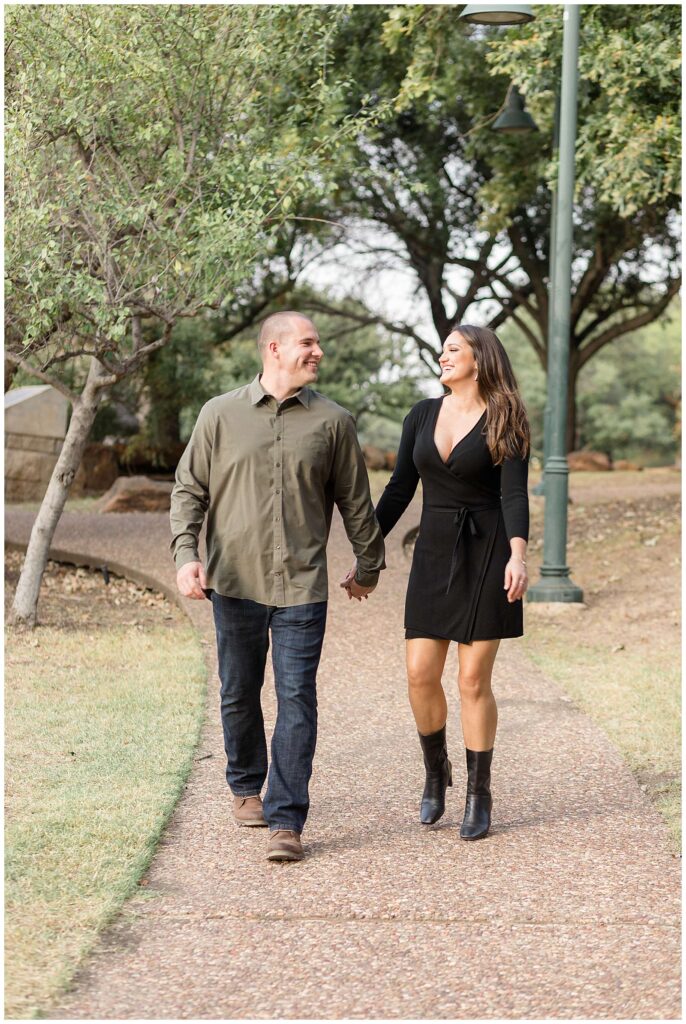 Couple walks down the path at Frisco Central Park holding hands and looking at each during Frisco mini sessions.  Woman wears a girl, cross, tie dress with black boots and he wears an olive button down and jeans.