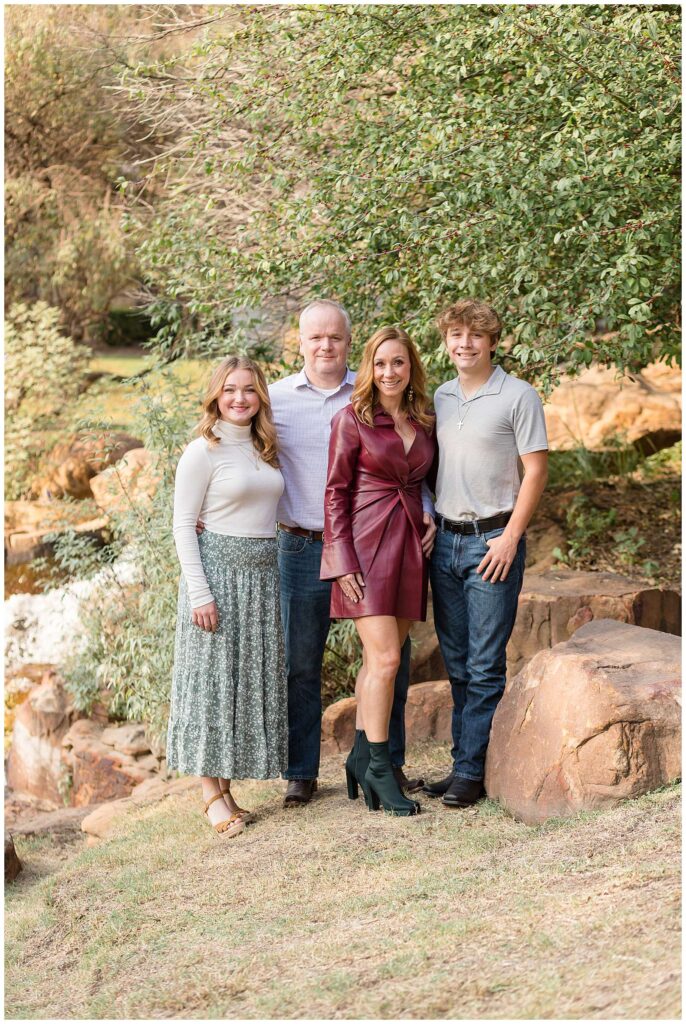 Family of four, with a teenage daughter and son, stand together and smile at the camera during their Frisco mini session.  Mom wears a burgundy leather dress with black booties, daughter wears a white turtleneck shirt and a green and white floral skirt, and Dad and son wear polo shirts and jeans.