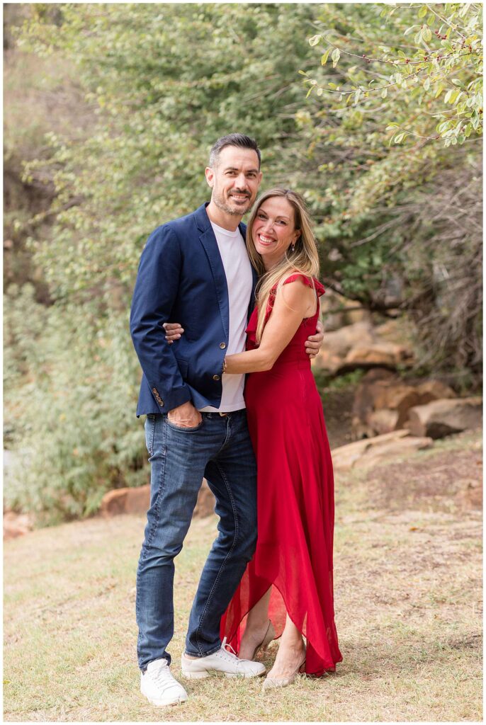 Couple stand together during their Frisco mini session with woman wearing a long, sleeveless, red dress and he wears jeans, a white shirt, and a blue blazer.  They stand close together and smile at the camera.
