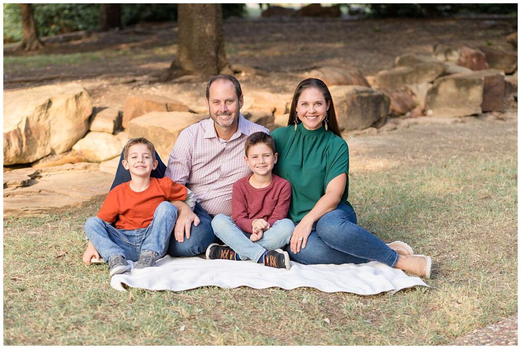 Family of 4, with two elementary aged sons, sit on a blanket on the grass and smile at the camera of Wisp + Willow Photography Co. during their Frisco mini session.