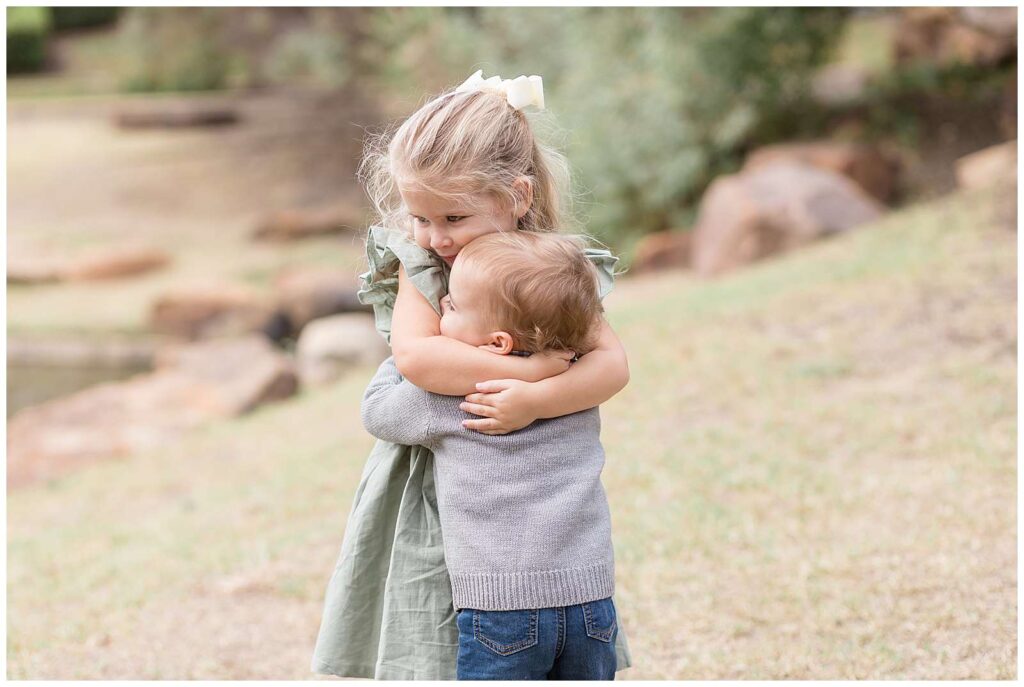 Sweet siblings, older, toddler sister hugs her younger, toddler brother as the hug each other tightly during their Frisco mini session.  THey both have blonde hair and cute toddler cheeks!