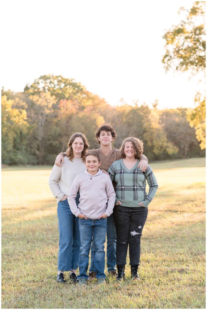 Chattanooga family photographer, Wisp + Willow Photography Co., take a picture of 4 kids as they stand together in the grass, surrounded by trees.  They all wear jeans and long sleeve shirts with fall leaves on some treets in the background.