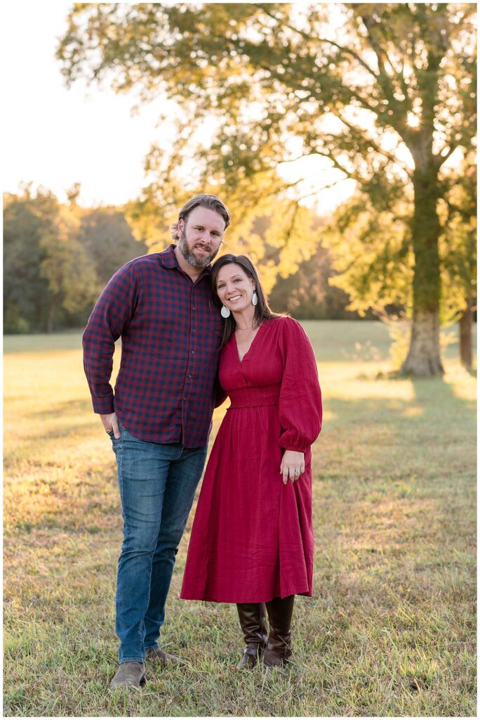 Chattanooga family photographer, Wisp + Willow Photography Co, capture a couple standing together with the wife's head on her husbands shoulder.  She wears a long red dress with white earrings and he wears a red and blue plaid button down shirt and jeans.