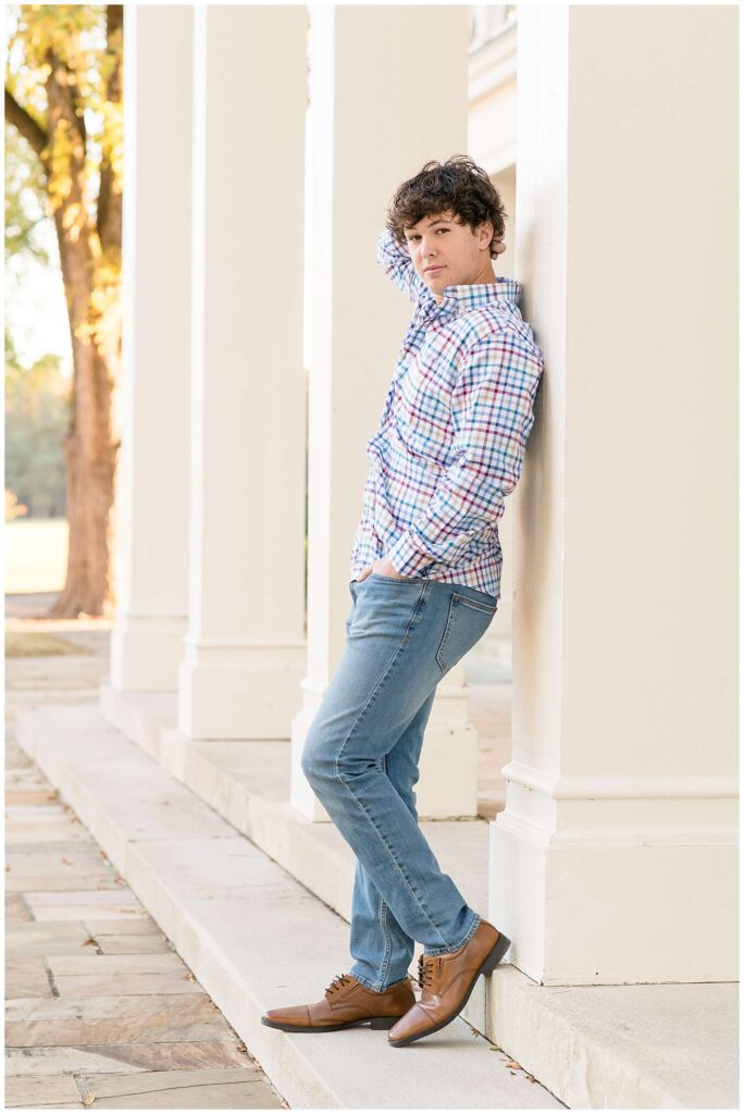 Senior boy stands against a white pillar with one hand in his pocket and the other behind his head at Chickamauga Battlefield in Chattanooga, TN wearing jeans, and a plaid, button down shirt.  