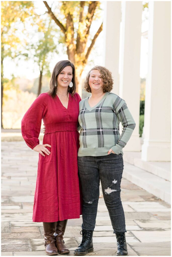 Mom and daughter stand together with their arms around each other and smiling at the camera of Wisp + Willow Photography Co.  Mom wears a red dress with white earrings and her daughter wears black jeans and a black and green plaid sweater.