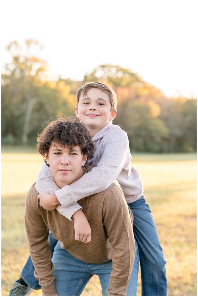 A blended Chattanooga family session shows an image of an older brother and little brother on his back as they both smile at the camera.  They wears jeans and coordinate in brown tone sweaters.