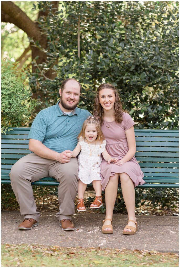 Family of 3, Mom, Dad, and toddler daughter sit on a park bench in Frisco Central Park in Frisco, TX for family portraits with Wisp + Willow Photography Co.  Check out the blog to see more!