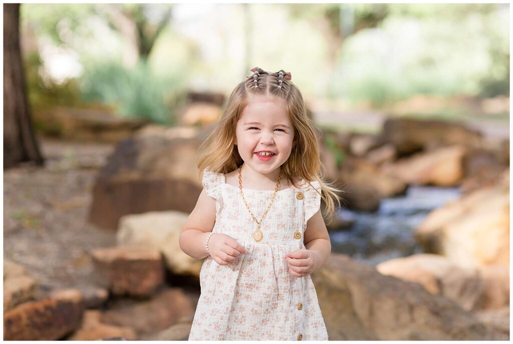 Toddler girl wears a gold necklace with a pink pendant, a white dress with floral pink flowers and two bubble braids on the top of her head and smiles big at the camera for her family portraits at Frisco Park.