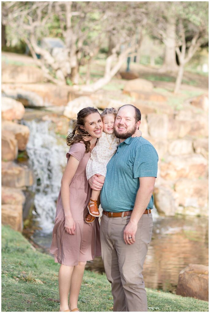 Frisco family portraits has Dad who wears a teal shirt and khaki pants, Mom who wears a mauve dress and their toddler daughter who they hold in-between them and hugs their necks, smiles at the camera of Wisp + Willow Photography Co.