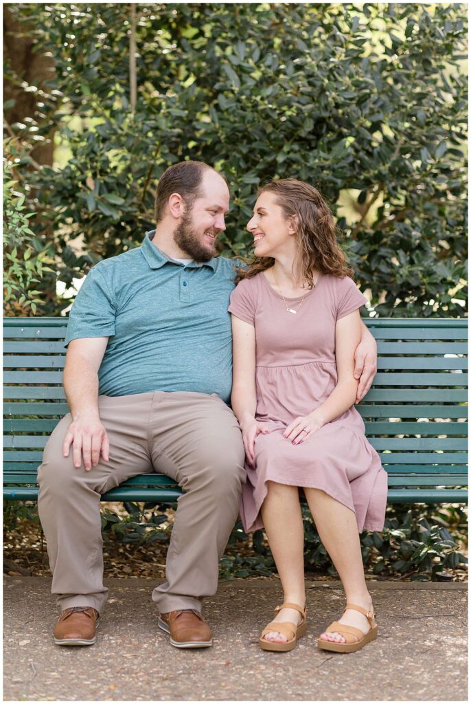 Couple sits on a park bench at Frisco Park and looks at each other and smiles during their Frisco family portraits.