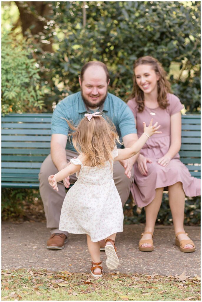 Mom and Dad sit on a park bench as their toddler daughter runs towards them and Dad reaches out his arms to catch her during their Frisco family portraits.