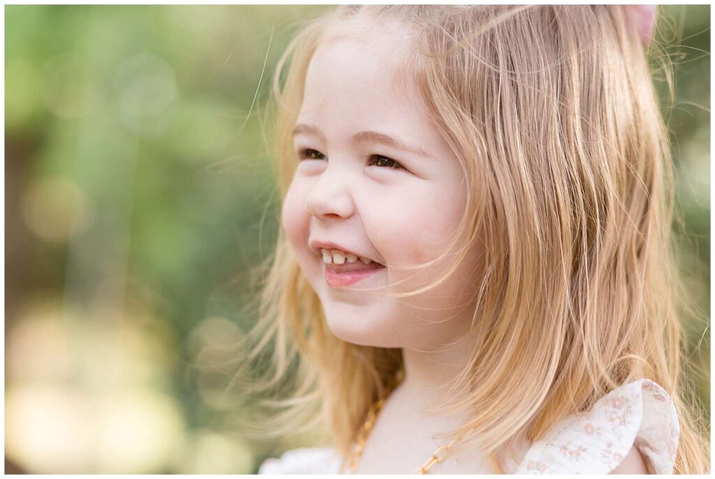 Photographer, Wisp + Willow Photography Co., capture a close up image of a toddler girl smiling in the distance with her sweet dimple, gold necklace, and white ruffle sleeve with pink flowers.