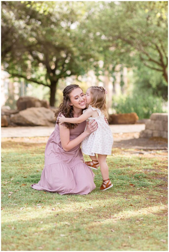 Mom, who is wearing a mauve dress bends down to hold her toddler daughter as she kisses her mom on the cheek during their Frisco family portraits at Frisco park.