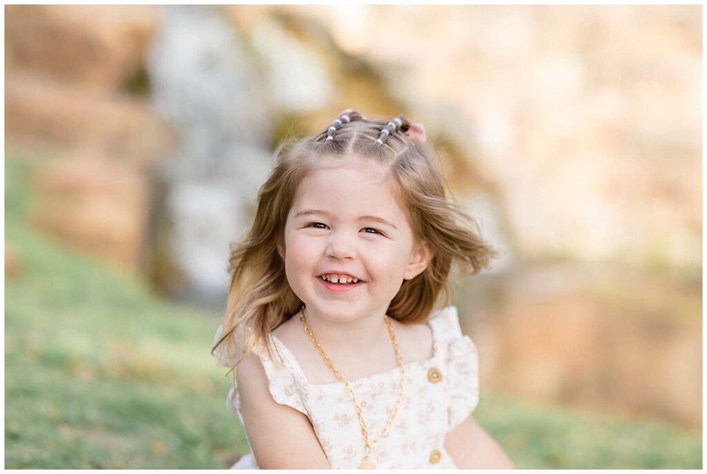 Texas photographer, Wisp + Willow Photography Co., take a picture of a toddler girl who smiles big at the camera wearing a gold necklace, white, ruffle sleeve dress with pink flowers, and two small bubble braids on the top of her head.