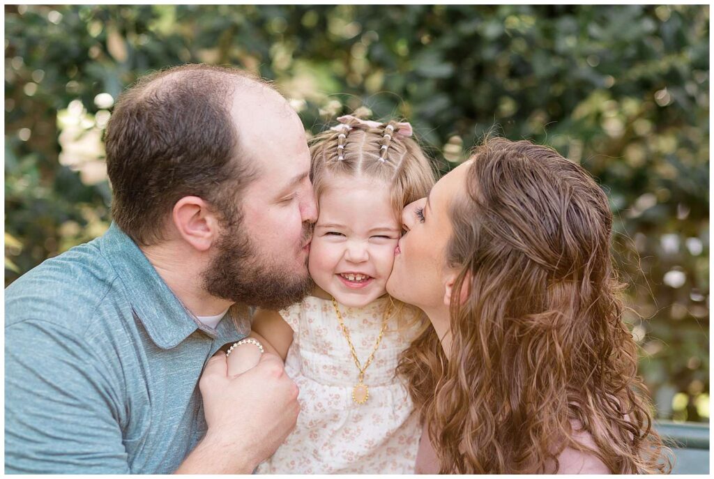 Mom and Dad give their toddler daughter big kisses on her cheeks as she smiles big at the camera during their Frisco family portrait session.
