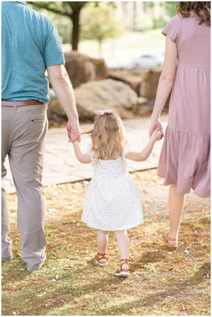 Texas photographer, Wisp + Willow Photography Co. capture a sweet image of a family of 3 walking away with the focus on their toddler daughter who holds her parents hands and wears a white dress, pink bow, and cognac, colored, strappy sandals.