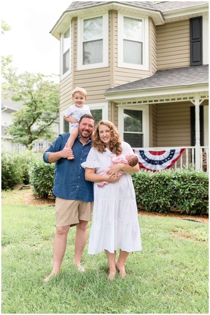 Family of 4, with a new big brother to his baby sister, stand in front of their Chattanooga home with an American flag hanging on their front porch banister.  They stand together as a new family of 4 for their lifestyle newborn session with Wisp + Willow Photography Co. 