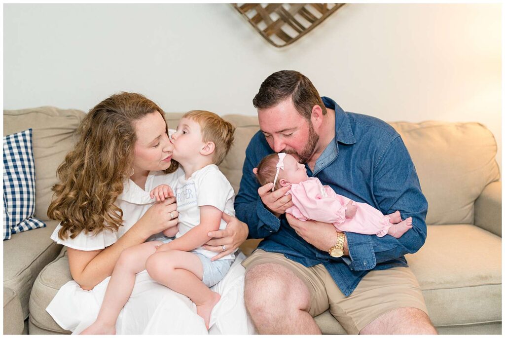 Family of 4 have a Chattanooga Lifestyle Newborn Session in their home with a new big brother and baby sister.  They sit on the coach together has their son gives his mom a kiss on her cheek and Dad gives his daughter a kiss on her forehead.