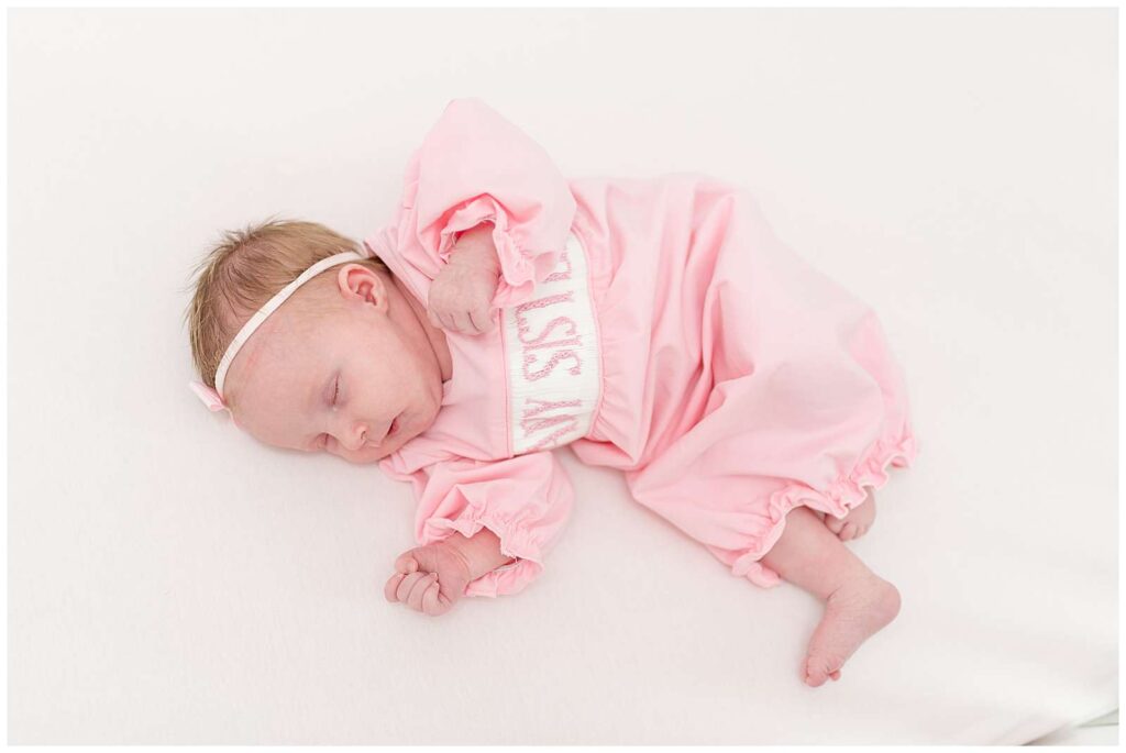 Newborn photographer, Wisp + Willow Photography Co, take a picture of a new baby girl who lays in her crib on a white sheet wearing a pink onesie with Baby Sister stitched on the front and she lays on her side.