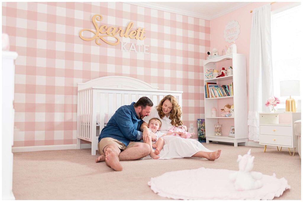New family of 4 sit on the floor in front of baby girl's crib and tickle one another.  The room is painted pink with one accent wall that has pink and white plaid wallpaper and Scarlett Kate woodwork on the wall.
