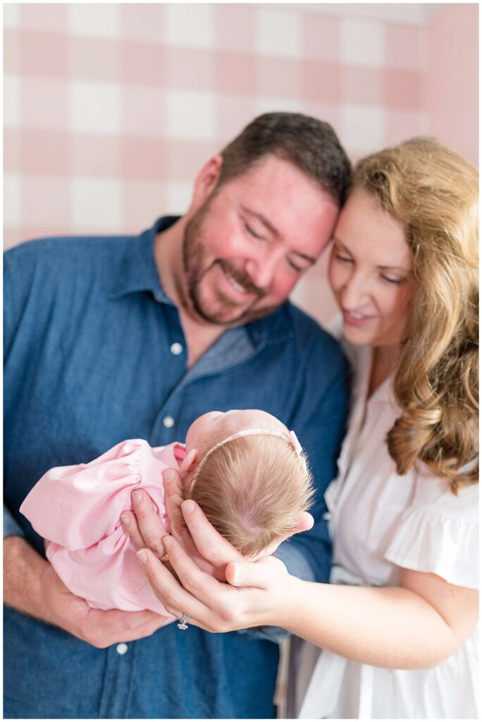 Newborn baby girl is held by Dad in his hands, with Mom also holding, as they look down at their baby girl. The Chattanooga Lifestyle Newborn Phototographer focuses on the baby's head full of hair.