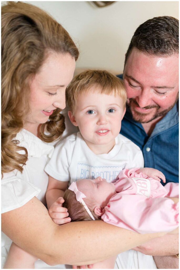 Chattanooga lifestyle newborn session takes a picture of a new family of 4 with Mom and Dad looking down at their baby girl in between them and their toddler son in the middle, smiles up at the camera holding his baby sister in front of him.