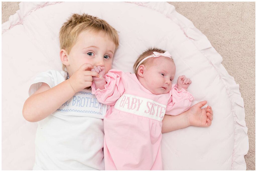 Big Brother and his new baby sister, lay on a round blanket on the floor and look up at newborn photographer, Wisp + Willow Photography Co.  They wear outfits that say Big Brother and Baby Sister stitched on the front.