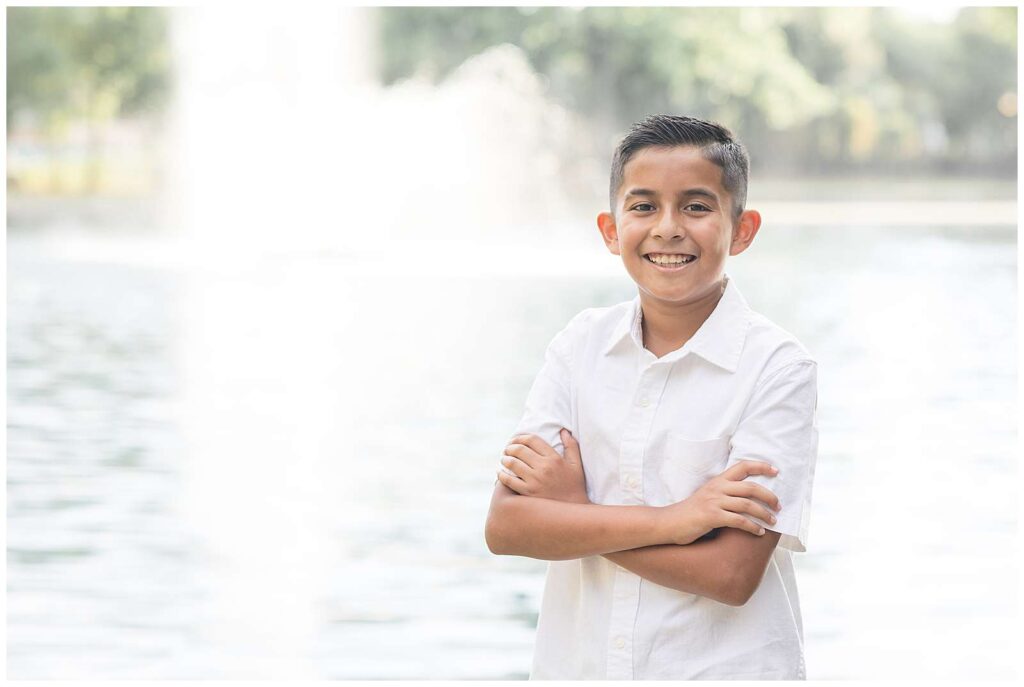 Oldest brother stands in front of water in Savannah, GA at Daffin Park with his arms crossed, wearing a white button shirt, and smiles at the camera.