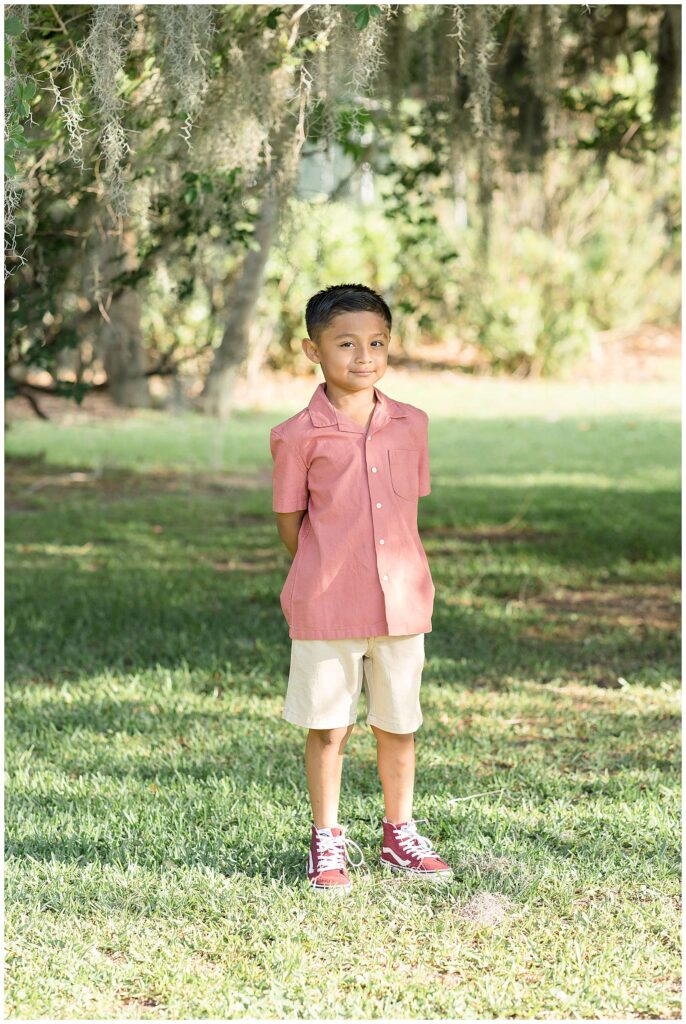 Little brother stands under a spanish moss tree wearing khaki shorts, a pink-peachish button down shirt and looks at the camera with his hands behind his back.