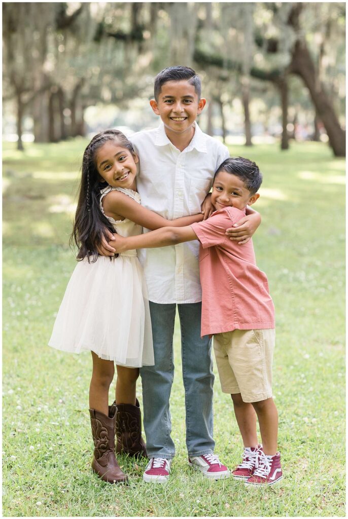 3 siblings wrap their arms around each other as they look at the camera of Wisp + Willow Photography Co.  The one sister and two brothers stand under the Spanish moss trees at Daffin Park.