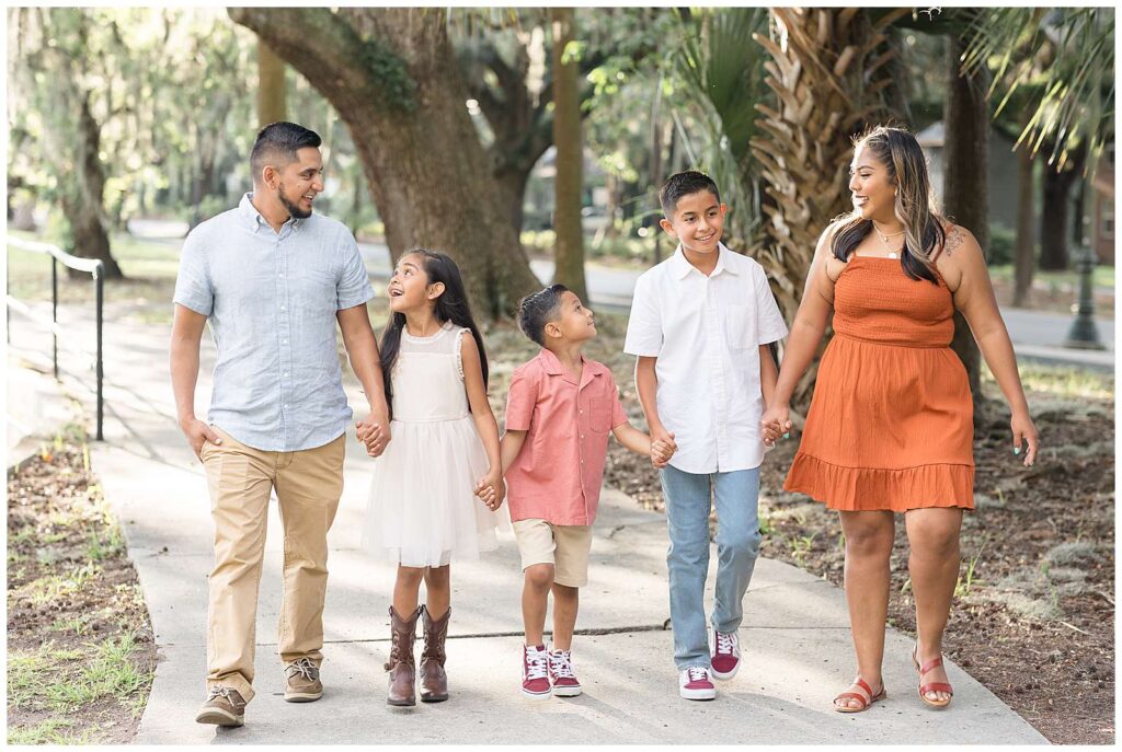 Family of 5 hold hands as they walk together with the parents on the ends and their one daughter and two sons in the middle.  They take Savannah family portraits in summer with Wisp + Willow Photography Co.