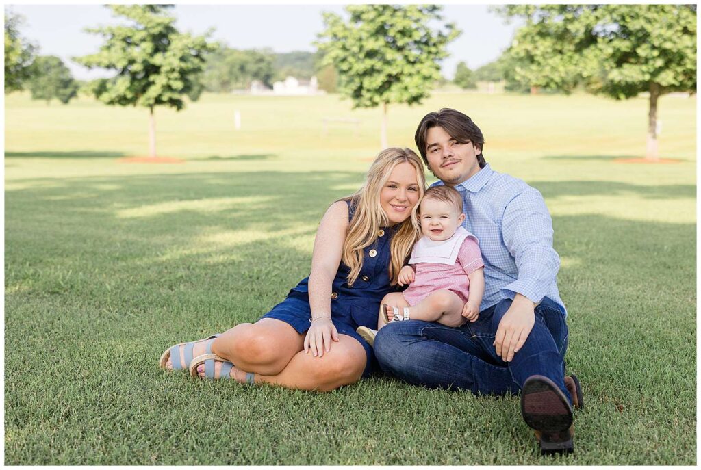 Family of 3 sit on the grass in Franklin, TN for their family portraits and their one year old son's cake smash session.  They coordinate in red, white, and blue and smile at the camera of Wisp + Willow Photography Co.