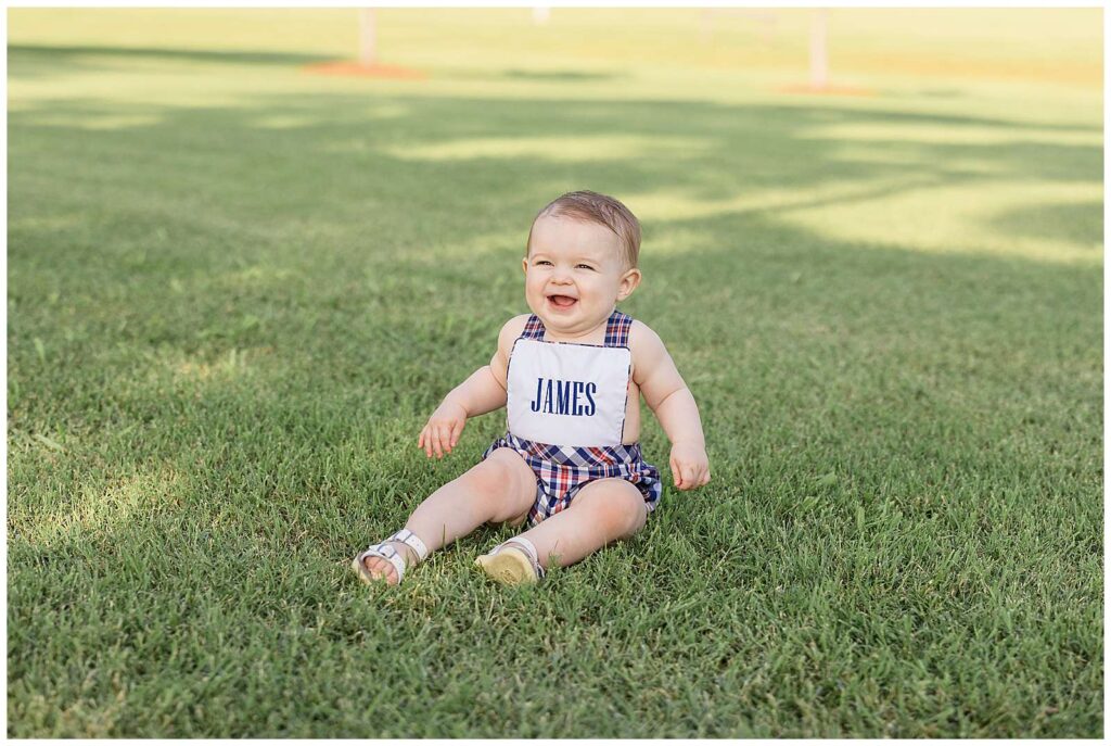 One year old boy sits on the grass in Franklin, TN wearing a red, white, and blue overall romper with his name on the front bib and he smiles at his parents just past the camera.