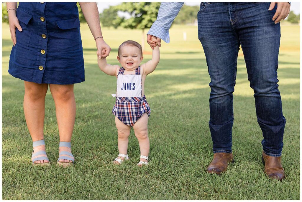 Sweet baby boy stands for his one year old portraits holding his Mom and Dad's hands and he wears a red,white, and blue overall romper and a monogrammed bib on the front with his name.