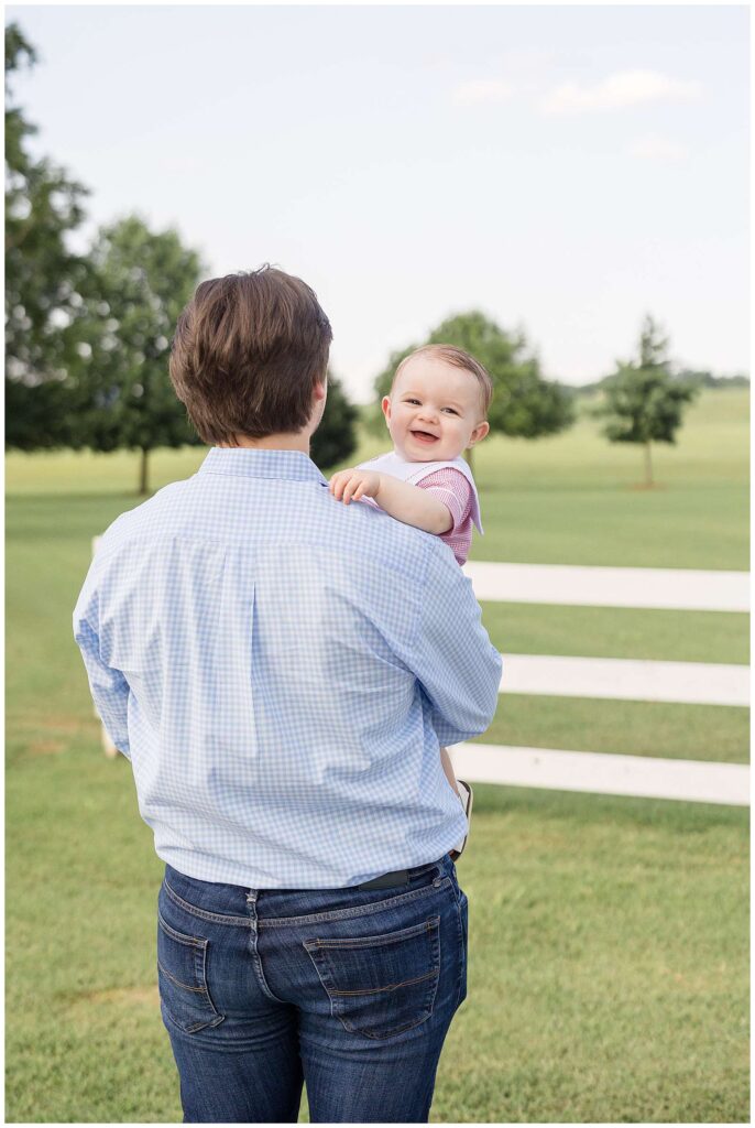 Dad stands facing away from the camera wearing jeans and a blue and white plaid shirt as he holds his son who looks over his shoulder and smiles back at the camera.