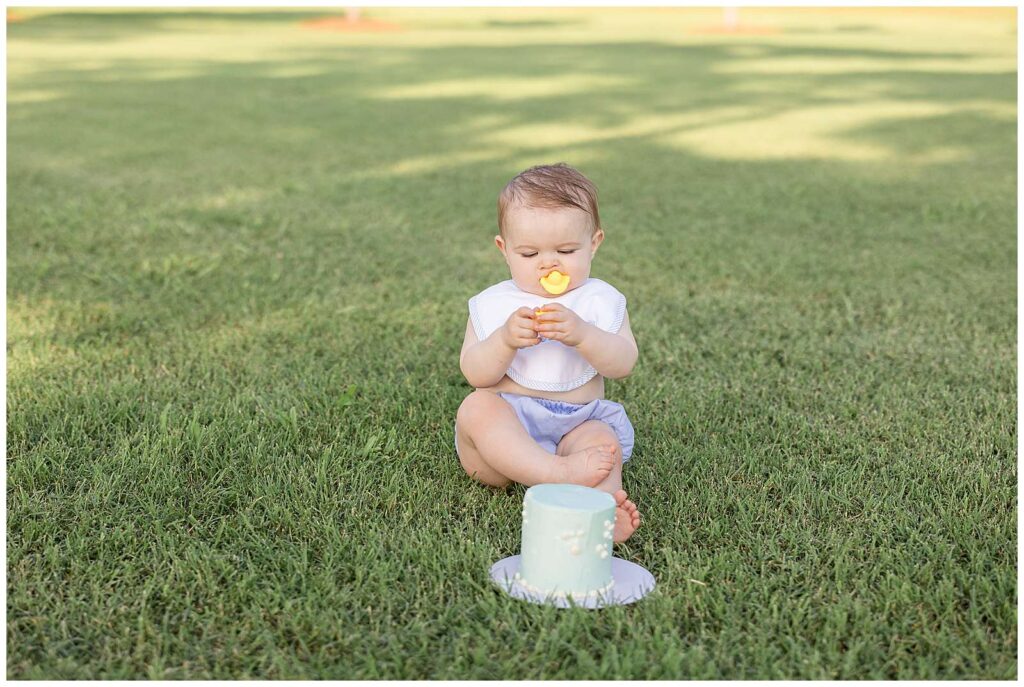 One year old boy sits just behind his smash cake on the grass being more interested in the rubber ducky toppers than the cake.  He wears a blue diaper cover, a bib, and puts one rubber ducky in his mouth and holds the other.