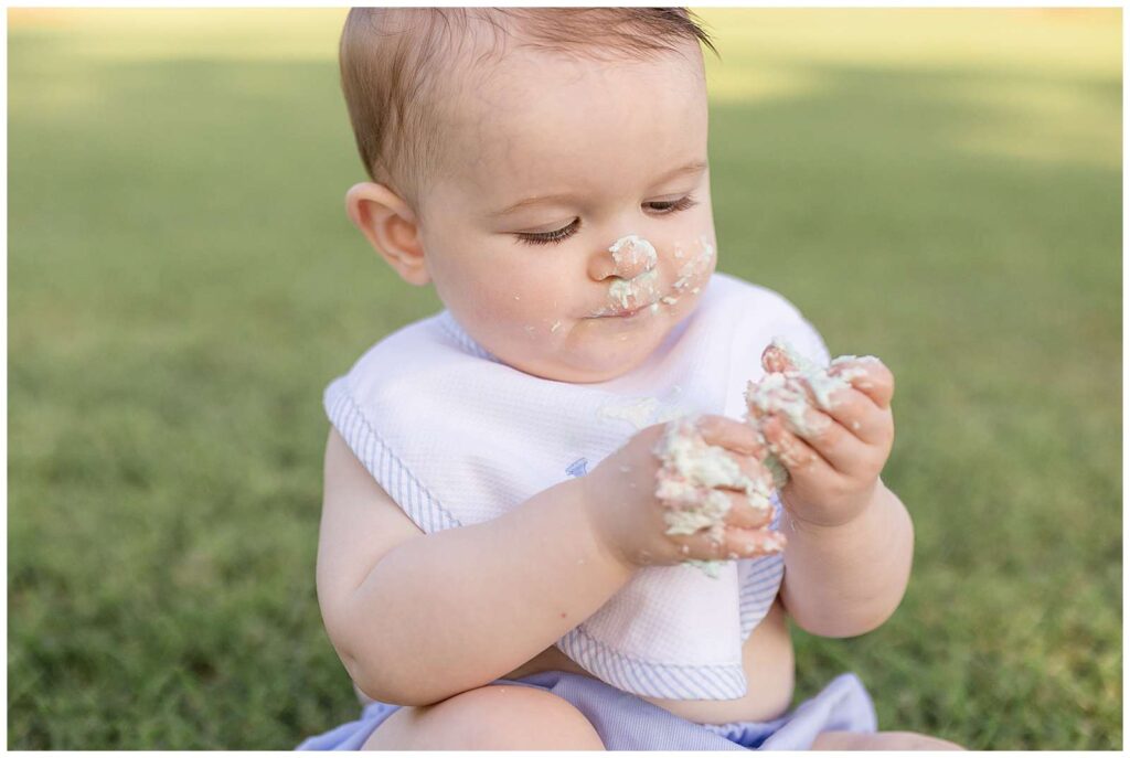 One year old boy sits in the grass with icing on his nose and face as he holds a piece of cake in his hands covered by icing.