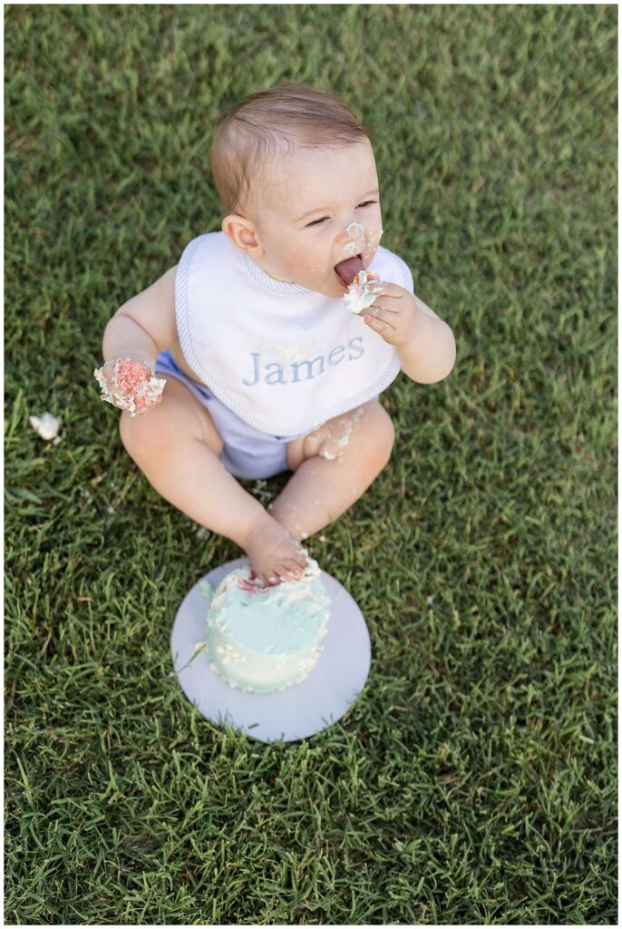 One year old baby boy sits on the grass with his feet crossed, toes in the icing on his smash cake, and cake being fisted in one hand and the other holding cake and being liked during this smash cake session in Franklin, TN.