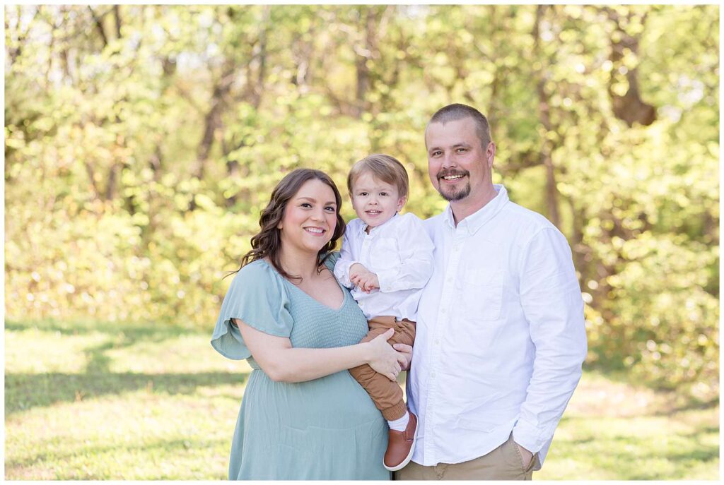 Family of 3 have a Texas maternity session at Erwin Park.  Dad and their toddler son both wears white button down shirts and khaki pants and Mom wears a muted teal dress with ruffle sleeves.