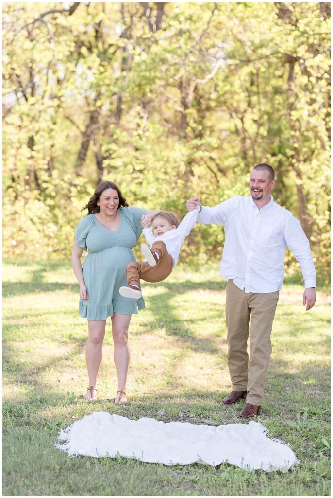 Texas maternity session at Erwin Park has Mom and Dad swinging their toddler son in the air and smiling big.  You can feel the joy in the image as the boys match with white button down shirts and khaki pants and Mom wears a short, teal, muted dress.