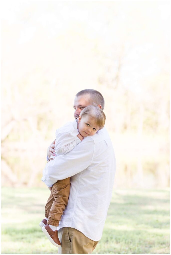 Toddler son gets tired at the end of his family session with Wisp + Willow Photography Co. and snuggles up to be held by Dad as he rests his tired head on his shoulder.   Dad and son match in their white button down shirts.