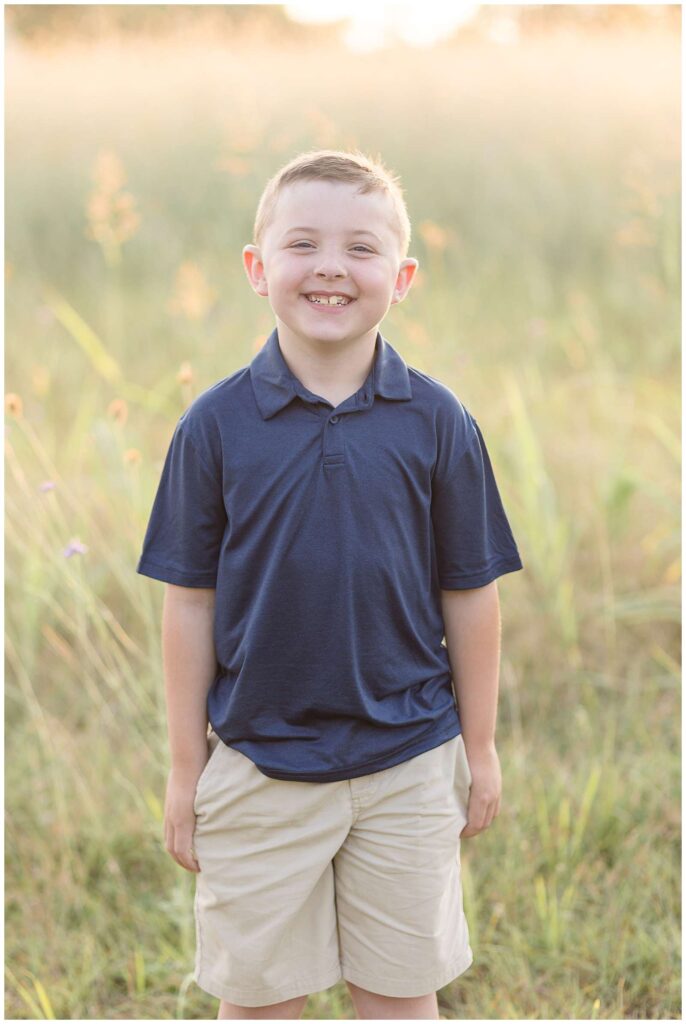 An elementary aged boy stands in the fields at Erwin Park in McKinney, TX during his family photography session with Wisp + Willow Photography Co and takes an individual portrait of him as he smiles at the camera.