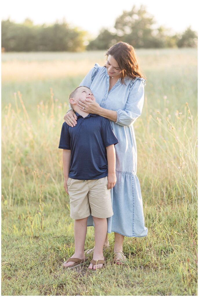 A mom stands behind her young son holding his face as he looks back at her and they smile at each other.  Mom wears a long, denim dress and her son wears a navy blue polo and khaki shorts for their Texas family photography session with photographer, Wisp + Willow Photography Co.