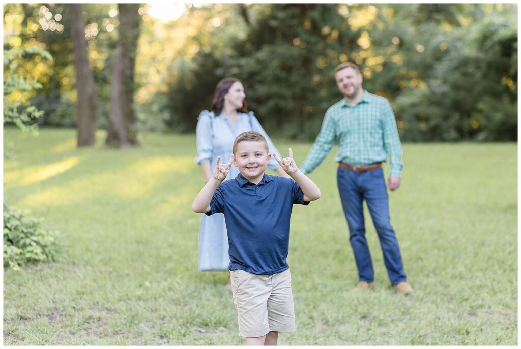 Elementary-aged boy stands in the forefront of an image with his parents in the background holding hands.  The boy wears a navy polo shirt and khakis and holds up his "rockstar" hands!